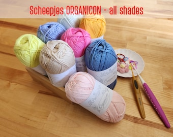 May include: A collection of Scheepjes Organicon yarn in various shades, including light blue, yellow, pink, beige, and light pink. The yarn is arranged in a wooden box with the label visible. There are also two crochet hooks and a small plate with crochet supplies.