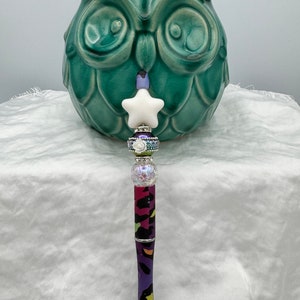 May include: A colorful pen with a purple, pink, and green animal print design. The pen has a white star charm and several clear and iridescent beads.