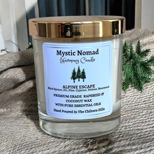 Puede incluir: Un tarro de vela de vidrio transparente con tapa dorada. La etiqueta dice "Mystic Nomad Harmony Candle" y "Alpine Escape". La vela está hecha con cera de colza y coco, y aceites esenciales puros. El texto también dice "Hand Poured In The Chiltern Hills."