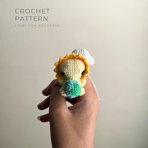 May include: A crocheted Chibi Sun keychain. The sun has a yellow body, a yellow and orange sunburst, and a small white cloud. The sun is holding a small blue and green ball. The text "CROCHET PATTERN CHIBI SUN KEYCHAIN" is at the top.