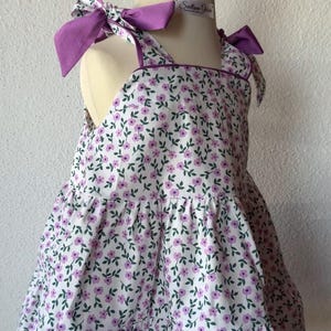 Può includere: Un vestito con stampa floreale bianca e viola con fiocchi viola sulle spalle. L'abito ha uno scollo quadrato e una gonna a balze. L'abito è esposto su un manichino.