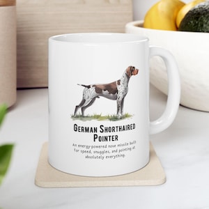May include: White ceramic mug featuring a German Shorthaired Pointer illustration. The dog is brown and white, standing on green grass. Text below reads "German Shorthaired Pointer" and a humorous description.