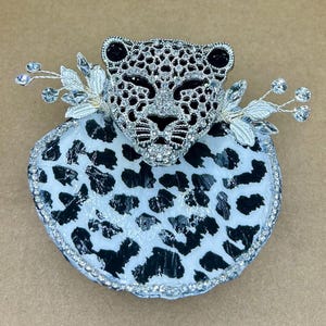 May include: A decorative brooch featuring a leopard head with a silver finish and sparkling rhinestones. The brooch has a black and white spotted base and is adorned with silver leaves and crystals.