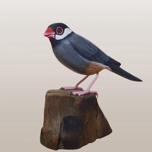 May include: A hand-carved wooden bird sculpture of a grey and white finch perched on a brown wooden stump. The bird has a red beak and pink feet.