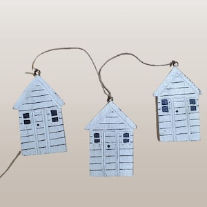 May include: Three white wooden beach hut decorations hanging from twine. Each hut has a black door and windows.