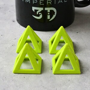 May include: Four lime green 3D printed pyramid-shaped objects with a hollow center. The pyramids are arranged in a square pattern on a gray surface. A black mug with the text "IMPERIAL 3D" is visible in the background.
