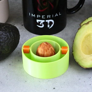 Puede incluir: Un pitter de aguacate verde con detalles naranjas, un aguacate entero y un aguacate cortado por la mitad. El pitter es una herramienta para quitar el hueso del aguacate.