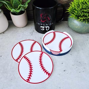 May include: Baseball-themed coasters with a white surface and red stitching detail. The coasters are round and have a red border. A black mug with the text "IMPERIAL 3D" is in the background, along with decorative plants.