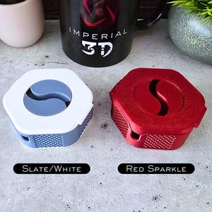 K&ouml;nnte beinhalten: Zwei 3D-gedruckte Kabelorganisatoren, einer in Schiefergrau und Wei&szlig; und der andere in Rot mit Glitzer. Die Organisatoren zeichnen sich durch ein einzigartiges Design mit einer geschwungenen Form und einer strukturierten Oberfl&auml;che aus.