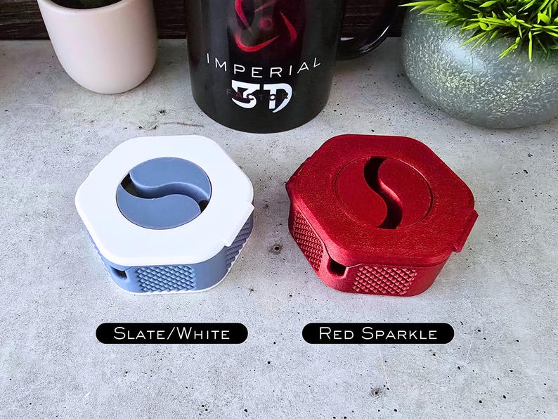 K&ouml;nnte beinhalten: Zwei 3D-gedruckte Kabelorganisatoren, einer in Schiefergrau und Wei&szlig; und der andere in Rot mit Glitzer. Die Organisatoren zeichnen sich durch ein einzigartiges Design mit einer geschwungenen Form und einer strukturierten Oberfl&auml;che aus.