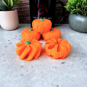 May include: Five orange pumpkin-shaped toys with green stems. The pumpkins are arranged in a pyramid shape on a gray surface.