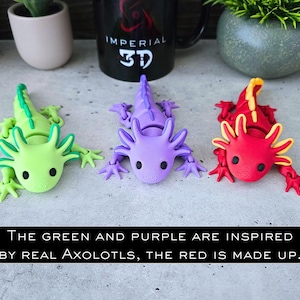 May include: Three articulated axolotl figures in bright green, purple, and red with yellow accents. Each figure features external gills, small black eyes, and a segmented body. Text on the image reads: The green and purple are inspired by real Axolotls, the red is made up.