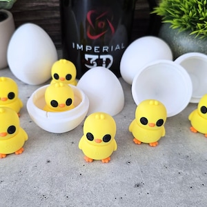 May include: A collection of yellow chick figurines with black eyes and orange feet. Some chicks are inside white eggshell halves, and others are standing. White egg-shaped containers are also present, along with a black container with white text.