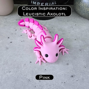 May include: A light pink articulated axolotl figurine with dark pink accents on the gills and tail fins. The figure features black eyes and a segmented body. Text above reads COLOR INSPIRATION: LEUCISTIC AXOLOTL and text below reads PINK.
