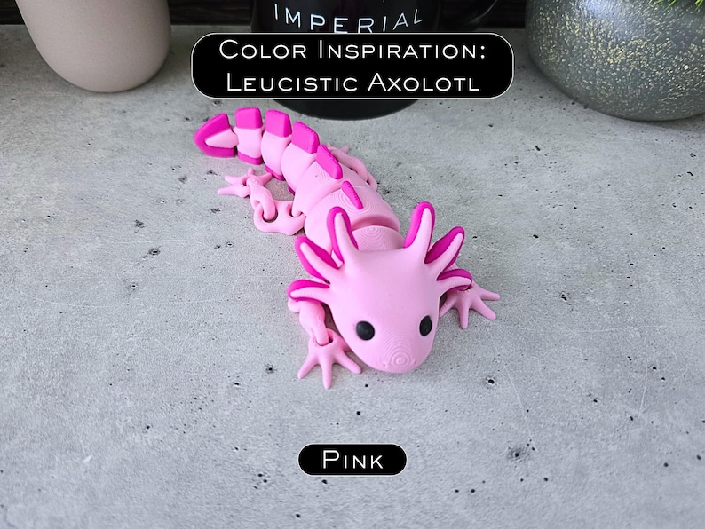 May include: A light pink articulated axolotl figurine with dark pink accents on the gills and tail fins. The figure features black eyes and a segmented body. Text above reads COLOR INSPIRATION: LEUCISTIC AXOLOTL and text below reads PINK.