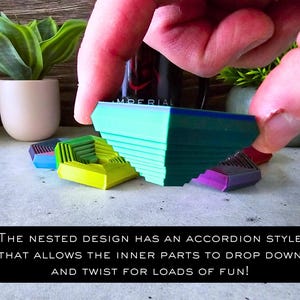 May include: A green, blue, and yellow geometric puzzle toy with a nested design. The toy is made of plastic and has an accordion-style mechanism that allows the inner parts to drop down and twist. The text "THE NESTED DESIGN HAS AN ACCORDION STYLE THAT ALLOWS THE INNER PARTS TO DROP DOWN AND TWIST FOR LOADS OF FUN!" is printed on a black background.