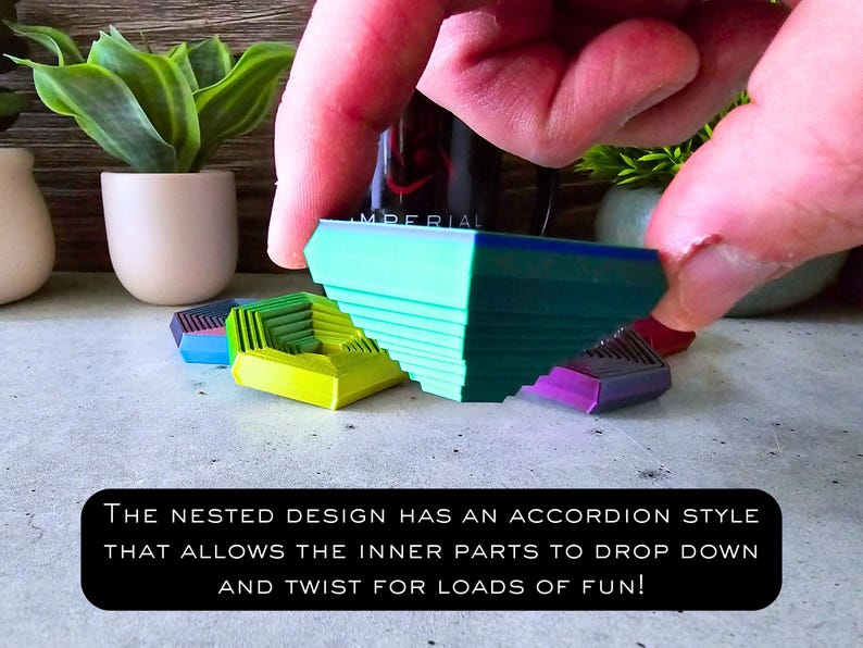 May include: A green, blue, and yellow geometric puzzle toy with a nested design. The toy is made of plastic and has an accordion-style mechanism that allows the inner parts to drop down and twist. The text "THE NESTED DESIGN HAS AN ACCORDION STYLE THAT ALLOWS THE INNER PARTS TO DROP DOWN AND TWIST FOR LOADS OF FUN!" is printed on a black background.