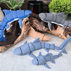 May include: A collection of articulated shark toys in various colors, including blue, gray, and tan. The sharks are arranged on a piece of driftwood, showcasing their segmented bodies and fins. The toys are designed with a playful aesthetic.