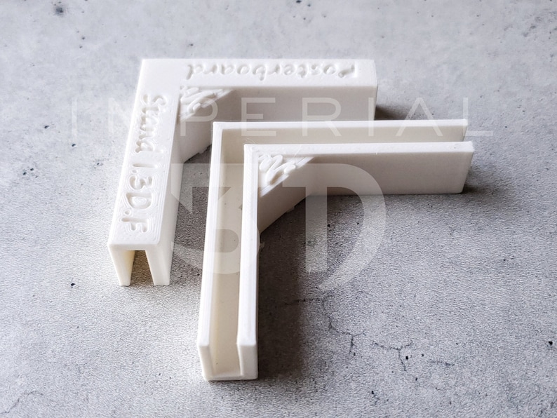 K&ouml;nnte beinhalten: Drei wei&szlig;e, L-f&ouml;rmige Posterboard-Halter mit dekorativen Eckakzenten. Die Texte "stand 1.3D.F" und "posterboard" sind sichtbar. Die Halter sind auf einer grauen Oberfl&auml;che angeordnet.