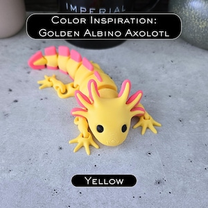 May include: A yellow and pink articulated axolotl figurine with black eyes. The text in the image reads: COLOR INSPIRATION: GOLDEN ALBINO AXOLOTL and YELLOW. The creature features a segmented body, four limbs, and external gills on the sides of its head.