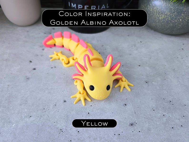 May include: A yellow and pink articulated axolotl figurine with black eyes. The text in the image reads: COLOR INSPIRATION: GOLDEN ALBINO AXOLOTL and YELLOW. The creature features a segmented body, four limbs, and external gills on the sides of its head.