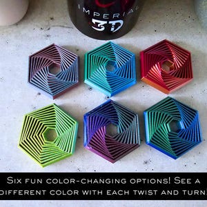 May include: Six hexagonal-shaped coasters with a color-changing design. Each coaster has a different color scheme, including blue, green, red, and purple. The coasters are made of a material that changes color when you twist or turn them. The text "IMPERIAL 3D" is visible on a black mug in the background.