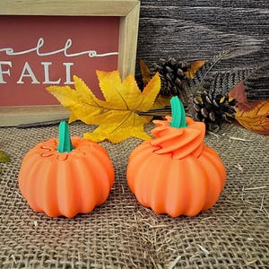 May include: Two orange pumpkins with green stems. One pumpkin has a standard stem, while the other features a swirled, textured top. The pumpkins are positioned in front of a wooden sign with the text "hello FALL" and autumn foliage.