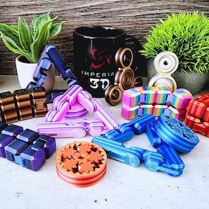 May include: A collection of colorful fidget toys, including a purple and pink triangle-shaped toy, a blue and white star-shaped toy, a rainbow cube, a brown and black cube, a blue and white fidget spinner, and a brown and orange gear-shaped toy. The toys are arranged on a wooden surface with a black mug that says "Imperial 3D" in the background.