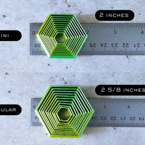 May include: Two green and black hexagonal cookie cutters, one measuring 2 inches and the other measuring 2 5/8 inches. The cookie cutters are made of metal and have a concentric circle design.