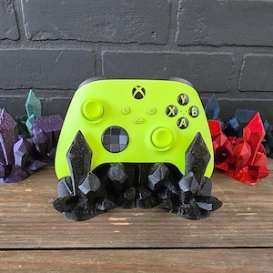 May include: A neon green Xbox controller resting on a black crystal-shaped stand. The stand is surrounded by decorative crystal formations in shades of green, purple, red, and blue. The controller has black buttons and the Xbox logo.