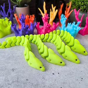 Op de afbeelding: Een limoengroen, beweeglijk aalspeelgoed met een gesegmenteerd lichaam en een gedetailleerde kop, liggend op een getextureerd oppervlak. Kleurrijke koraalvormige decoraties in blauw, paars, oranje, roze en geel zijn op de achtergrond.