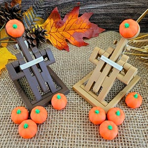 Puede incluir: Dos catapultas de madera, una marrón y otra beige, con caramelos naranjas con forma de calabaza. Las catapultas están decoradas con una banda blanca. Hojas de otoño y heno en el fondo.
