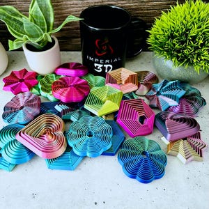 Puede incluir: Una colección de 15 formas geométricas coloridas impresas en 3D. Las formas están hechas de plástico en capas y tienen un acabado arcoíris iridiscente. Las formas incluyen un corazón, una estrella, una flor, un hexágono y un pentágono. Las formas están dispuestas sobre una superficie blanca. El texto "IMPERIAL 3D" es visible en una taza negra en el fondo.