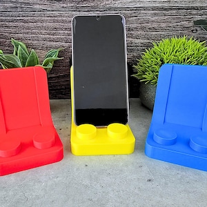May include: Three phone stands made to look like Lego bricks. The stands are red, yellow, and blue. The yellow stand has a black phone in it.
