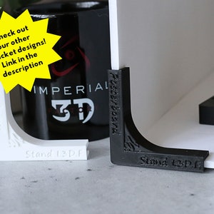 May include: Two 3D printed corner brackets, one white and one black, with the text "Stand 1.3D.F" printed on each. The brackets are designed to support a poster or other flat surface.