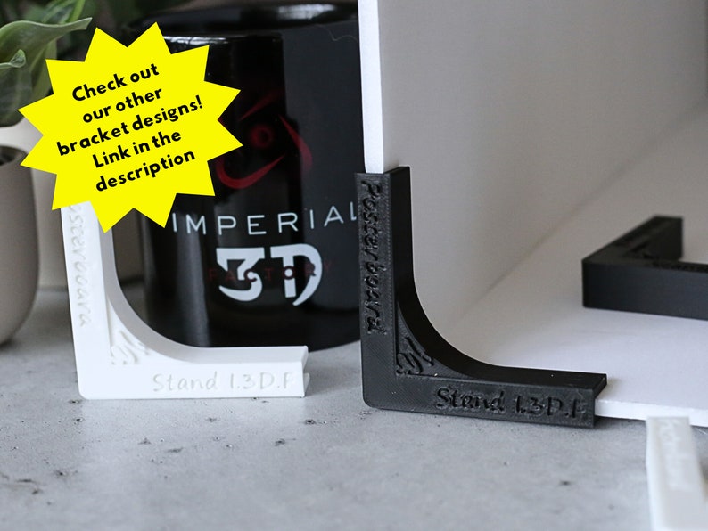 May include: Two 3D printed corner brackets, one white and one black, with the text "Stand 1.3D.F" printed on each. The brackets are designed to support a poster or other flat surface.