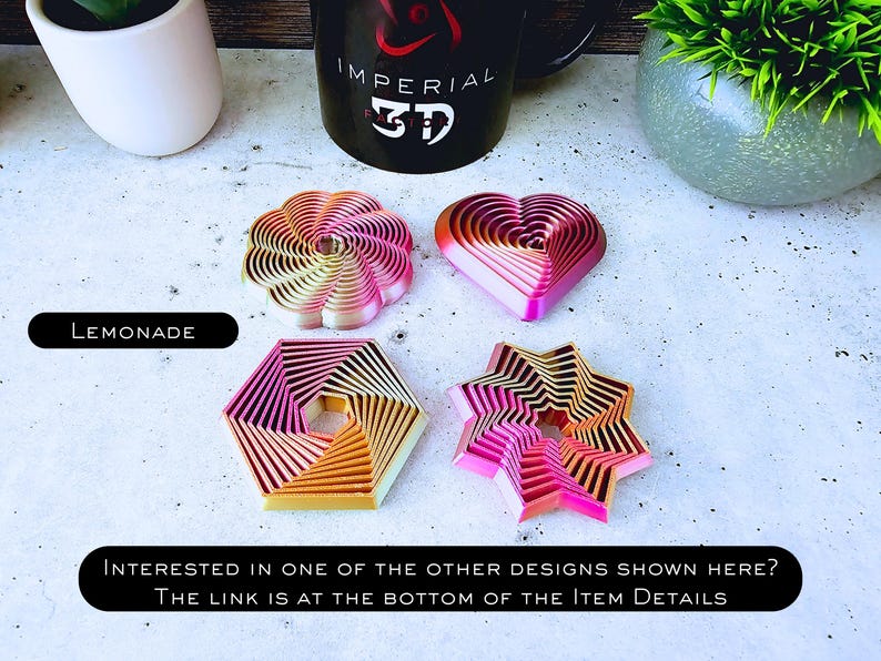May include: Five different cookie cutters in various shapes, including a heart, a star, a flower, and two geometric shapes. The cookie cutters are made of metal and have a pink and orange ombre finish. The text "LEMONADE" is visible at the top of the image.
