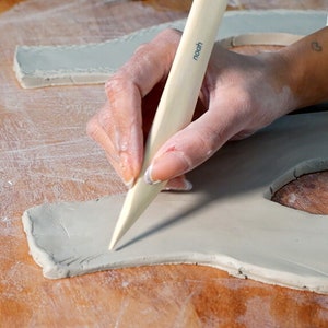 May include: A person using a wooden sculpting tool to shape a piece of gray clay. The tool has the word "noodh" printed on it.