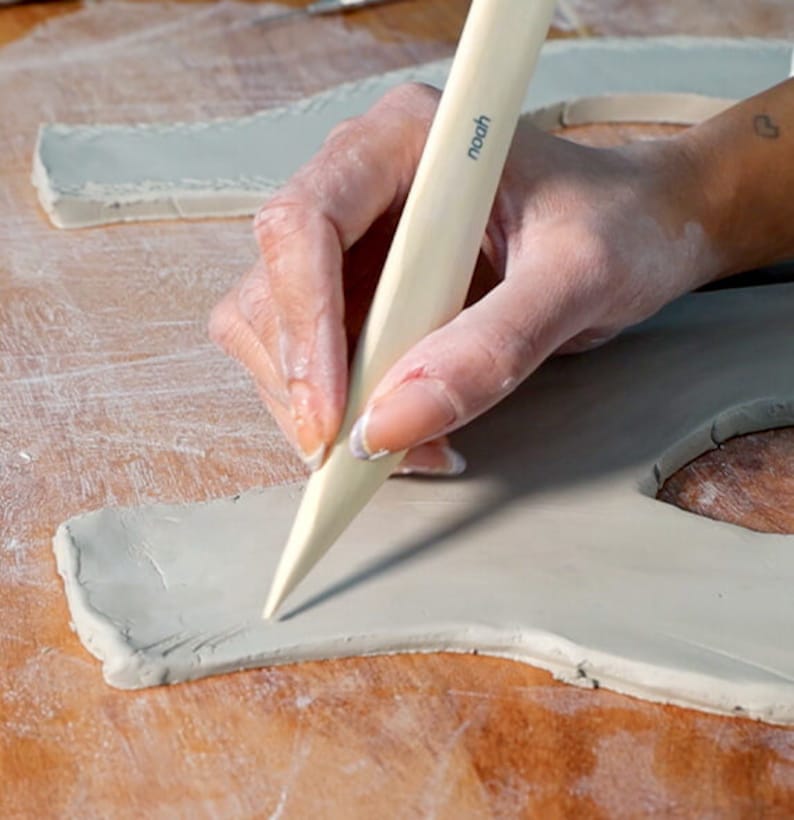 May include: A person using a wooden sculpting tool to shape a piece of gray clay. The tool has the word "noodh" printed on it.