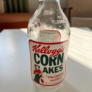 Op de afbeelding: Een doorzichtige glazen melkfles met een rood en groen Kellogg's Corn Flakes-etiket. Het etiket toont een haan en de tekst "The Original and Best".
