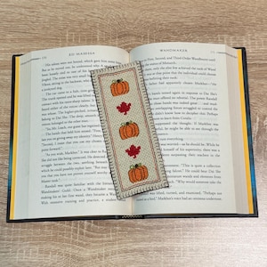 May include: A cross-stitched bookmark with a design of three orange pumpkins and two red maple leaves. The bookmark has a beige background and a dark border, ideal for marking pages in a book during autumn.