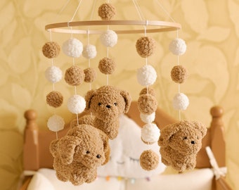 May include: A wooden baby mobile with brown and white pom-poms and crocheted brown teddy bears hanging from it. The mobile is hanging above a wooden crib with white bedding.