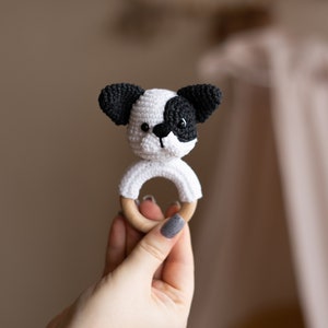 May include: A crocheted dog toy with black ears and a white face, attached to a wooden ring. The toy is being held by a hand.