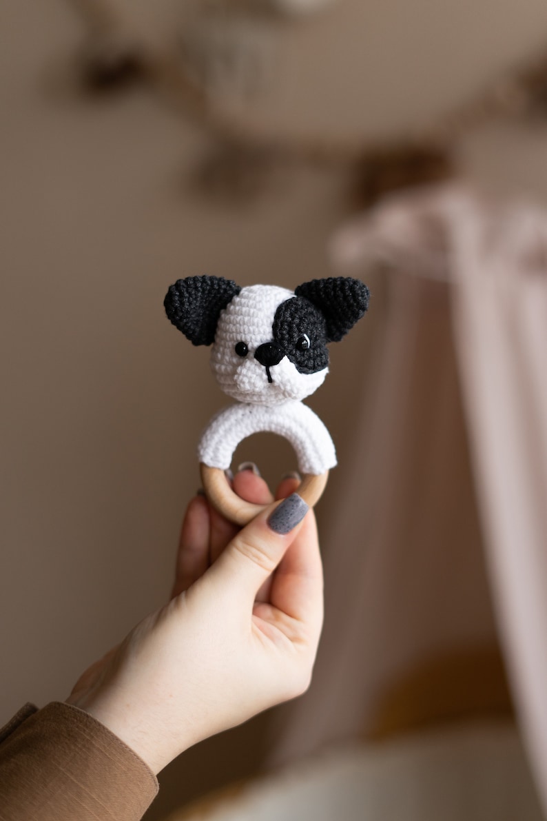 May include: A crocheted dog toy with black ears and a white face, attached to a wooden ring. The toy is being held by a hand.