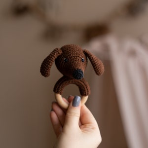 May include: A brown crocheted dog head attached to a wooden teething ring. The ring is held in a hand with gray nail polish.