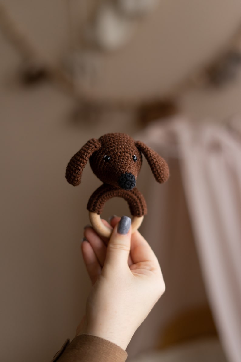 May include: A brown crocheted dog head attached to a wooden teething ring. The ring is held in a hand with gray nail polish.