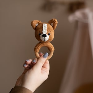 May include: A crocheted brown and white dog rattle with a wooden ring. The rattle is held in a hand with gray nail polish.