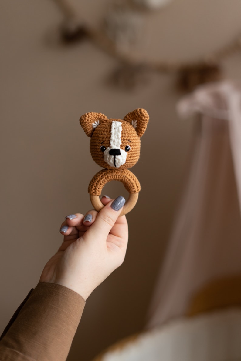 May include: A crocheted brown and white dog rattle with a wooden ring. The rattle is held in a hand with gray nail polish.