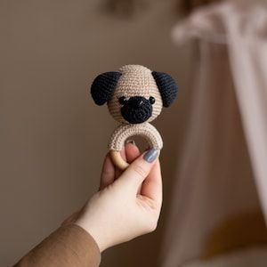 May include: A crocheted beige pug dog with black ears and nose attached to a wooden ring. The ring is held in a hand.