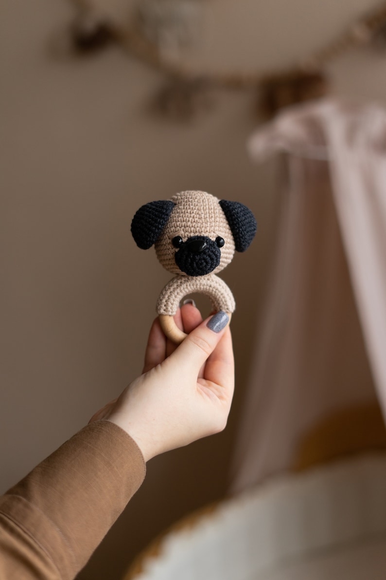 May include: A crocheted beige pug dog with black ears and nose attached to a wooden ring. The ring is held in a hand.
